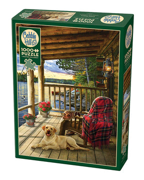Puzzle Cobble Hill Cabin Porch – 1000 pièces. Scène paisible montrant une véranda en bois au bord d’un lac avec un chien allongé, une chaise avec couverture tartan, une lanterne suspendue, le tout sur carton rigide avec découpe aléatoire, finition lin. Illustration créée par Greg Giordano, dimensions assemblée env. 67,5 × 48,8 cm.